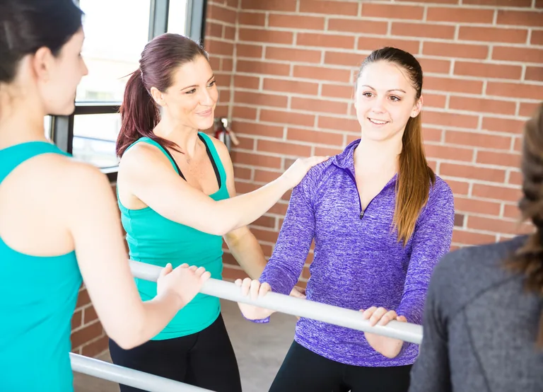 Two participants at a studio barre, with one guiding the other at the shoulder.