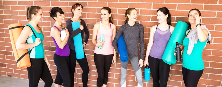 A group talks in a bright studio while holding exercise mats and water bottles.